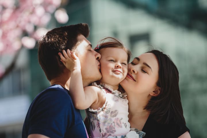 Unos padres jóvenes besan a una niña en la mejilla desde ambos lados bajo un árbol rosa en flor, el apego con los padres es un factor de protección de la resiliencia