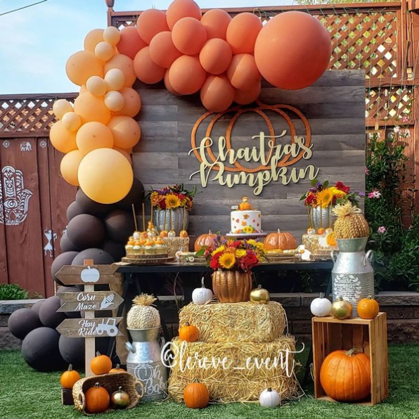 bebé en otoño con una mesa de golosinas rodeada de globos naranjas y marrones, heno y calabazas con un fondo de madera 