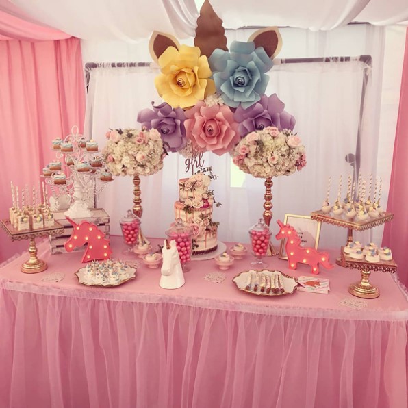 baby shower de unicornio con una mesa rosa que tiene accesorios para el unicornio y flores de colores