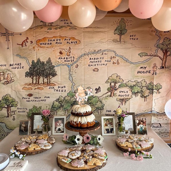 Baby shower de Winnie the Pooh con un mapa de un libro rodeado de globos pastel rosas, blancos y naranjas y una mesa de postres